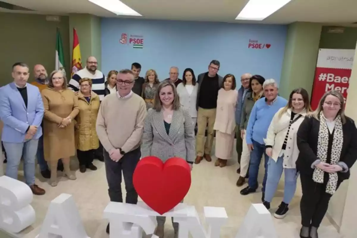 Un grupo de personas posando en una sala con letras grandes que forman la palabra Baena y un corazón rojo, al fondo hay banderas y logotipos del PSOE en la pared