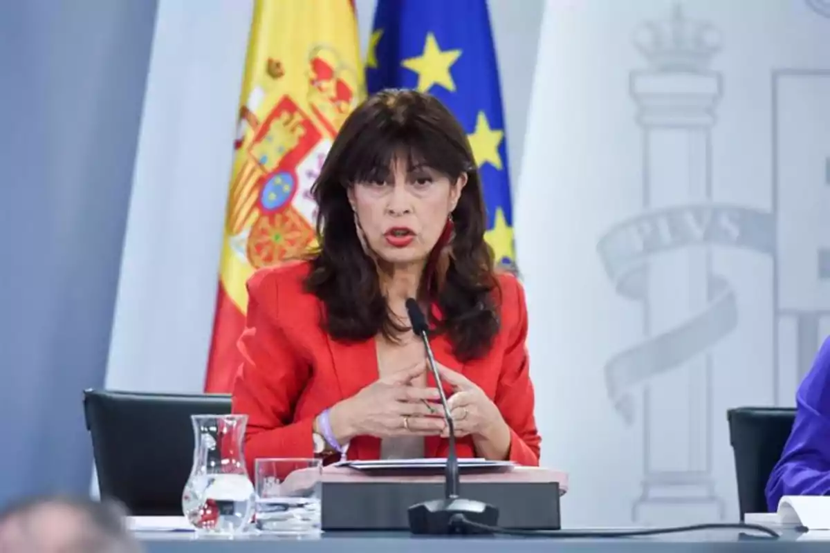Mujer de cabello oscuro con chaqueta roja hablando frente a un micrófono en una conferencia con banderas de España y la Unión Europea al fondo Mujer de cabello oscuro con chaqueta roja hablando frente a un micrófono en una conferencia con banderas de España y la Unión Europea al fondo