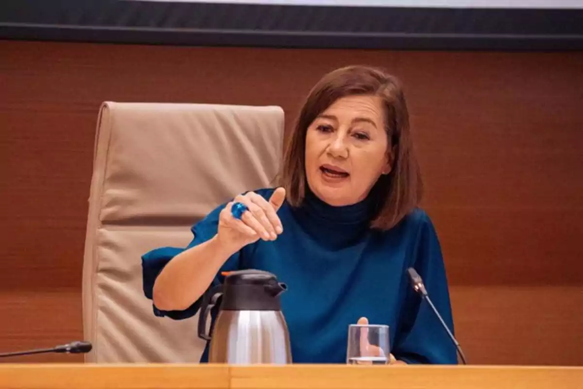 Mujer sentada en una silla grande hablando en una mesa con un micrófono, una jarra plateada y un vaso de agua frente a ella Mujer sentada en una silla grande hablando en una mesa con un micrófono, una jarra plateada y un vaso de agua frente a ella