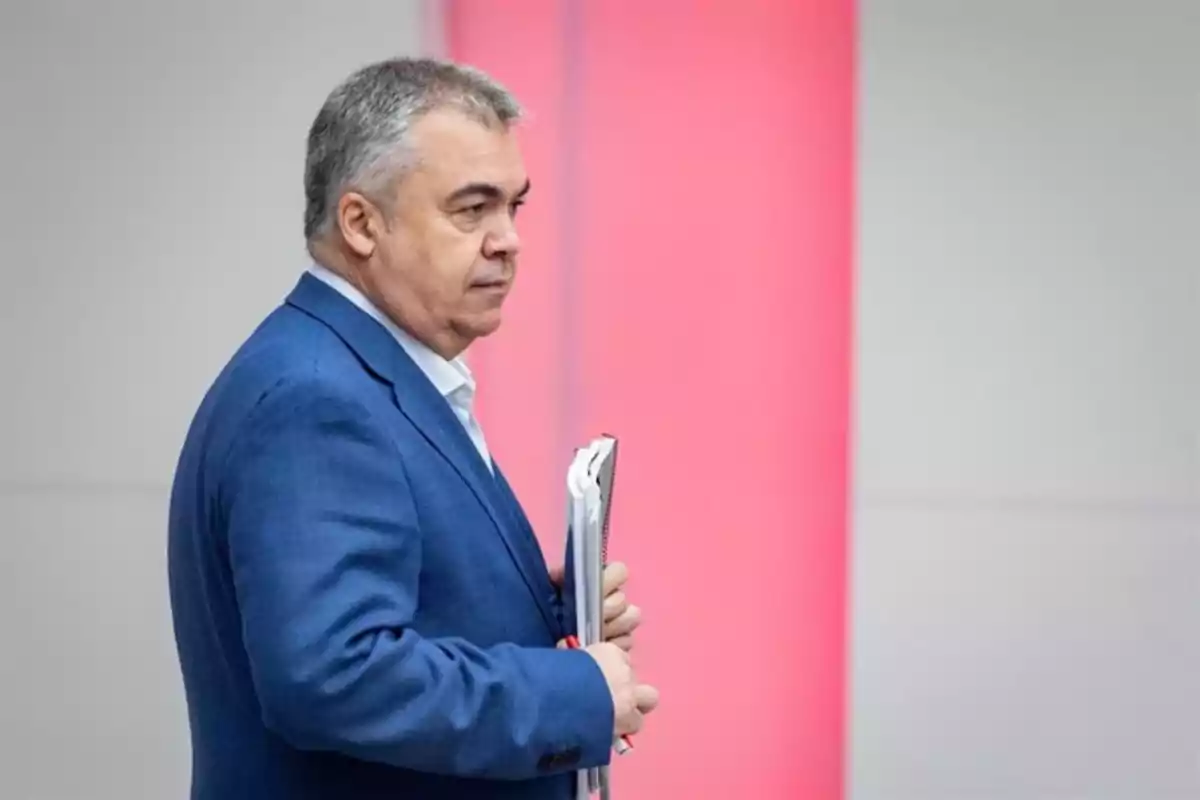 Hombre de cabello canoso con saco azul sosteniendo carpetas y documentos frente a una pared con fondo rosa y gris. Hombre de cabello canoso con saco azul sosteniendo carpetas y documentos frente a una pared con fondo rosa y gris.