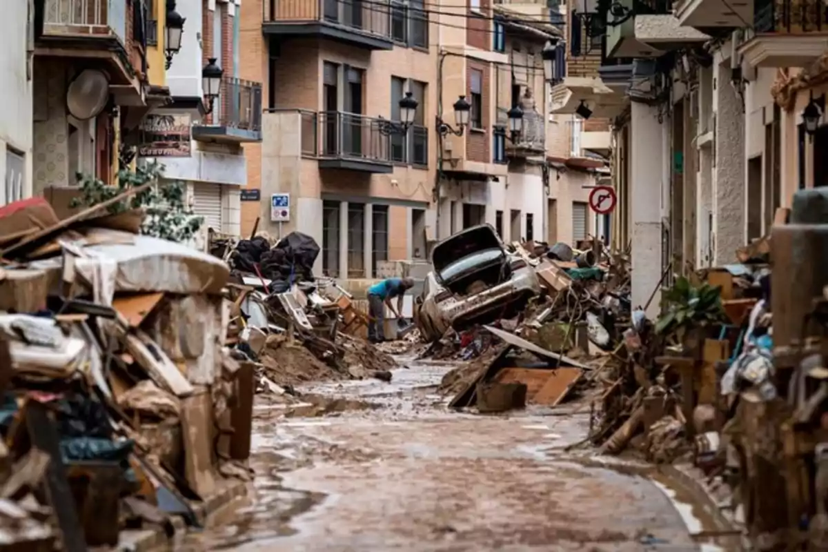 Calle de una ciudad llena de escombros, muebles y un coche dañado tras una inundación, con una persona limpiando entre los restos Calle de una ciudad llena de escombros, muebles y un coche dañado tras una inundación, con una persona limpiando entre los restos