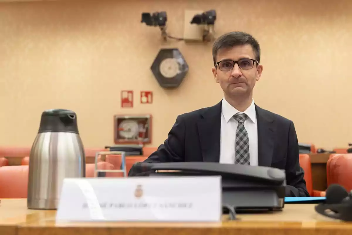 Hombre con gafas y traje sentado en una sala de reuniones con una cafetera de acero inoxidable y un cartel con su nombre sobre la mesa Hombre con gafas y traje sentado en una sala de reuniones con una cafetera de acero inoxidable y un cartel con su nombre sobre la mesa