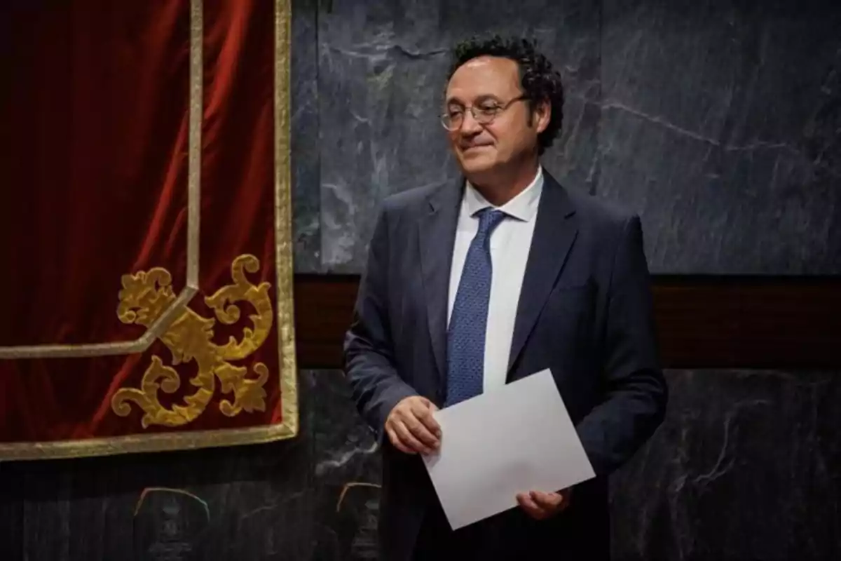 Hombre de cabello rizado y gafas con traje azul y corbata sosteniendo una hoja en las manos de pie frente a una pared de mármol y una cortina roja decorada con detalles dorados Hombre de cabello rizado y gafas con traje azul y corbata sosteniendo una hoja en las manos de pie frente a una pared de mármol y una cortina roja decorada con detalles dorados