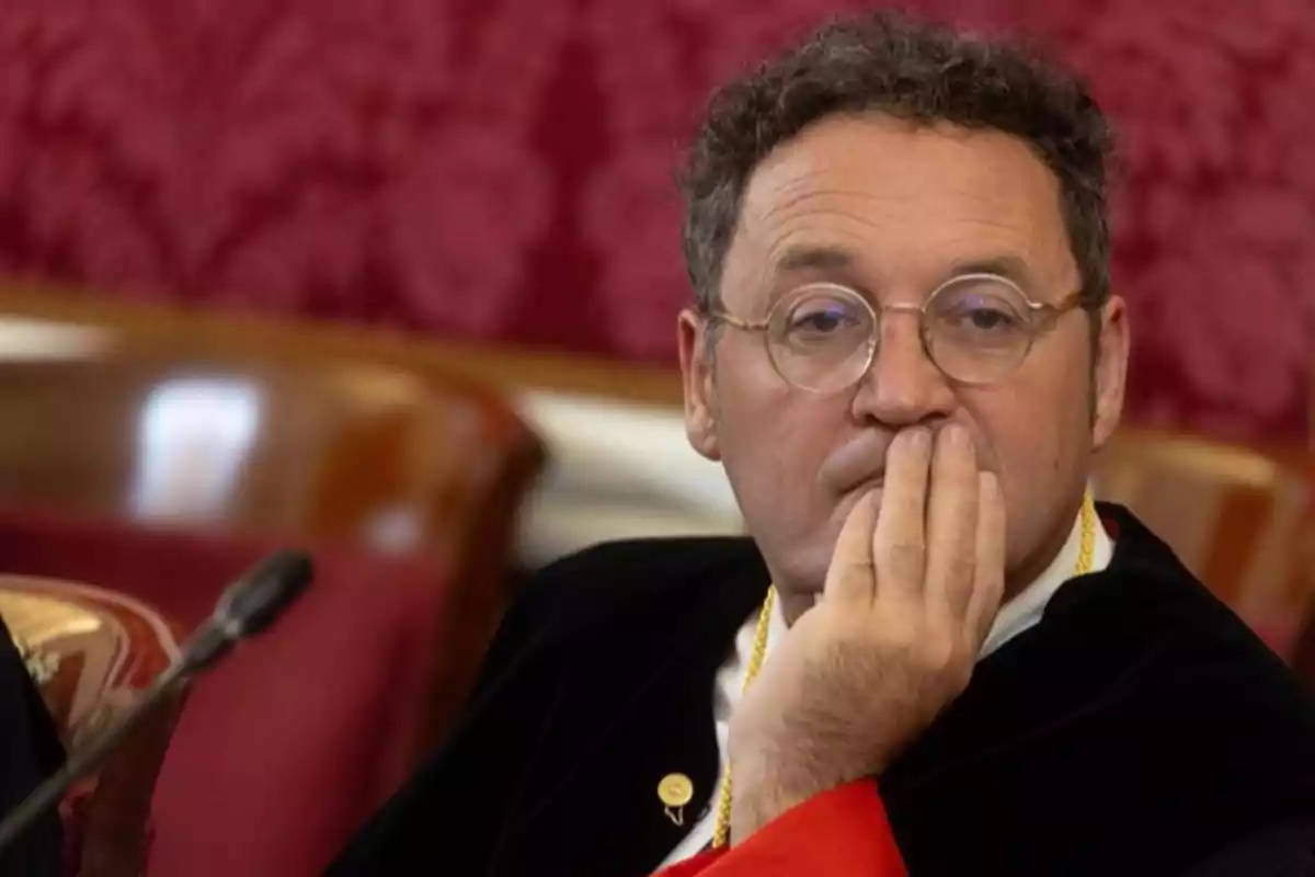 Hombre con gafas y toga negra sentado en una sala elegante con fondo rojo, apoyando la mano en su rostro y expresión pensativa Hombre con gafas y toga negra sentado en una sala elegante con fondo rojo, apoyando la mano en su rostro y expresión pensativa
