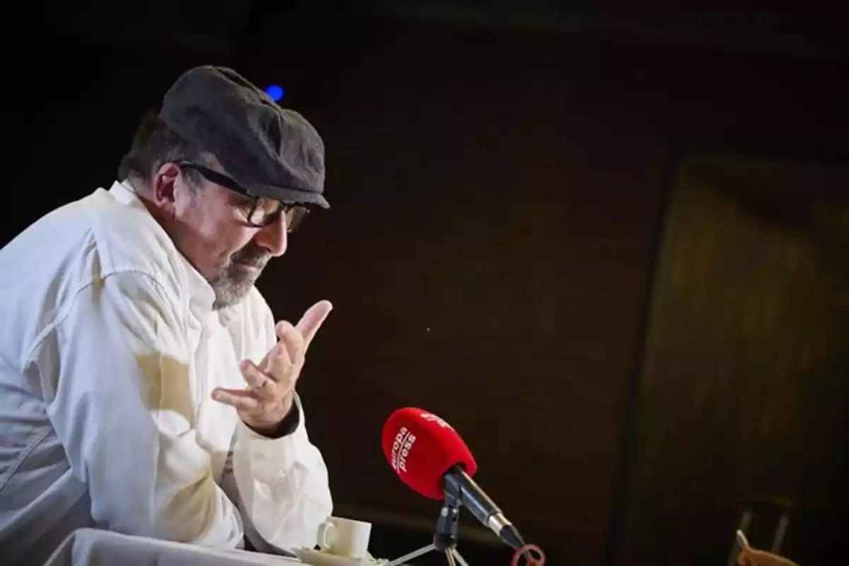 Hombre con gafas y gorra hablando frente a un micrófono rojo en una mesa con una taza de café Hombre con gafas y gorra hablando frente a un micrófono rojo en una mesa con una taza de café