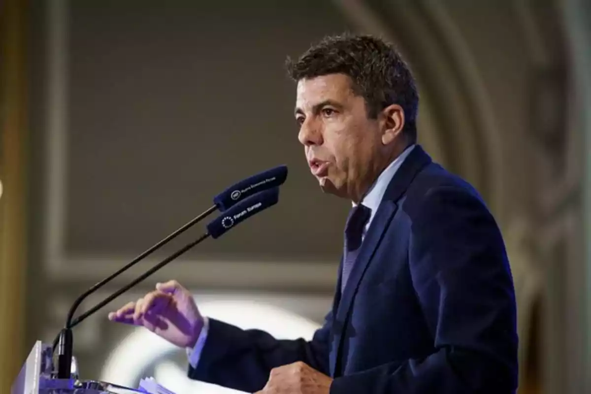 Hombre de traje azul hablando en un podio con dos micrófonos en un entorno formal Hombre de traje azul hablando en un podio con dos micrófonos en un entorno formal