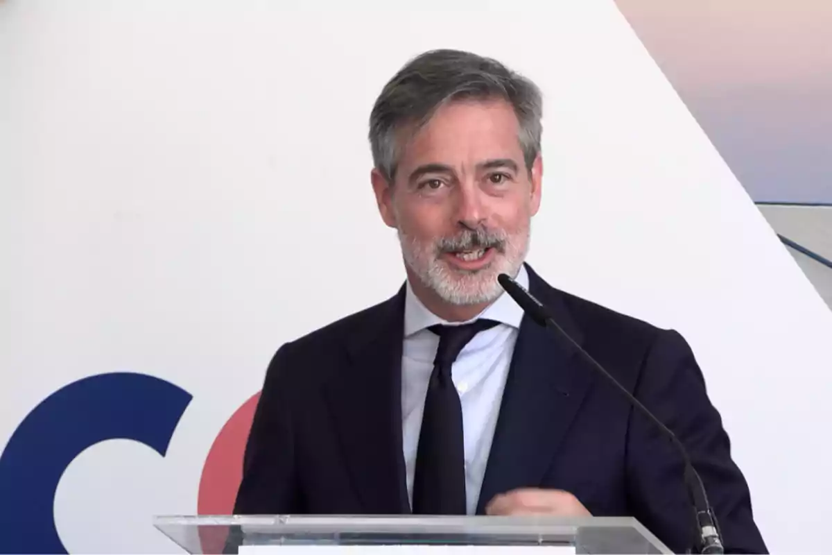 Hombre de cabello canoso y barba hablando en un podio con micrófono, usando traje oscuro y corbata, fondo claro con formas geométricas. Hombre de cabello canoso y barba hablando en un podio con micrófono, usando traje oscuro y corbata, fondo claro con formas geométricas.