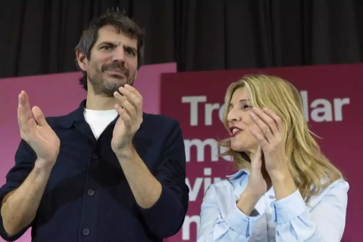 Un hombre y una mujer aplauden juntos en un escenario con fondo rosa y letras blancas. Un hombre y una mujer aplauden juntos en un escenario con fondo rosa y letras blancas.