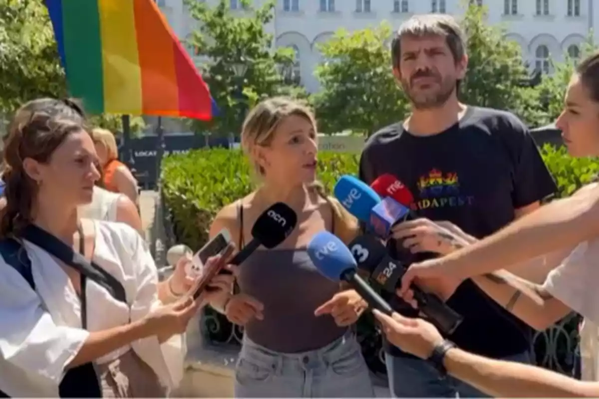 Varias personas dan declaraciones a la prensa al aire libre mientras una bandera arcoíris ondea en el fondo y varios micrófonos de diferentes medios rodean a la mujer que habla. Varias personas dan declaraciones a la prensa al aire libre mientras una bandera arcoíris ondea en el fondo y varios micrófonos de diferentes medios rodean a la mujer que habla.