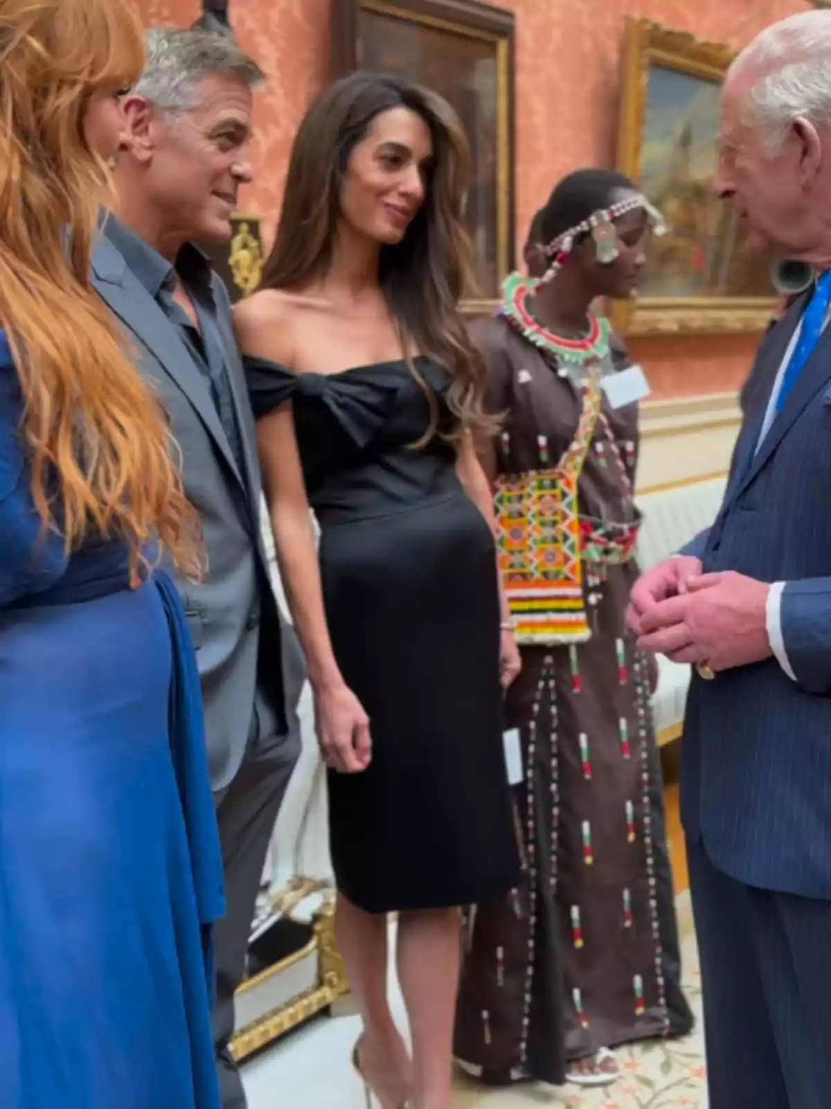 Varias personas elegantemente vestidas conversan en una sala decorada con cuadros y muebles clásicos, una de ellas lleva un vestido tradicional con cuentas de colores. Varias personas elegantemente vestidas conversan en una sala decorada con cuadros y muebles clásicos, una de ellas lleva un vestido tradicional con cuentas de colores.