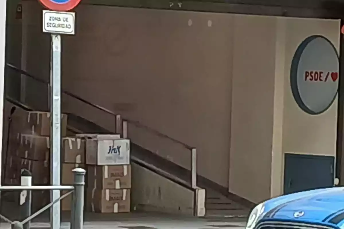 Varios cartones apilados junto a una rampa en la entrada de un edificio con un cartel de PSOE y un coche azul estacionado en primer plano Varios cartones apilados junto a una rampa en la entrada de un edificio con un cartel de PSOE y un coche azul estacionado en primer plano