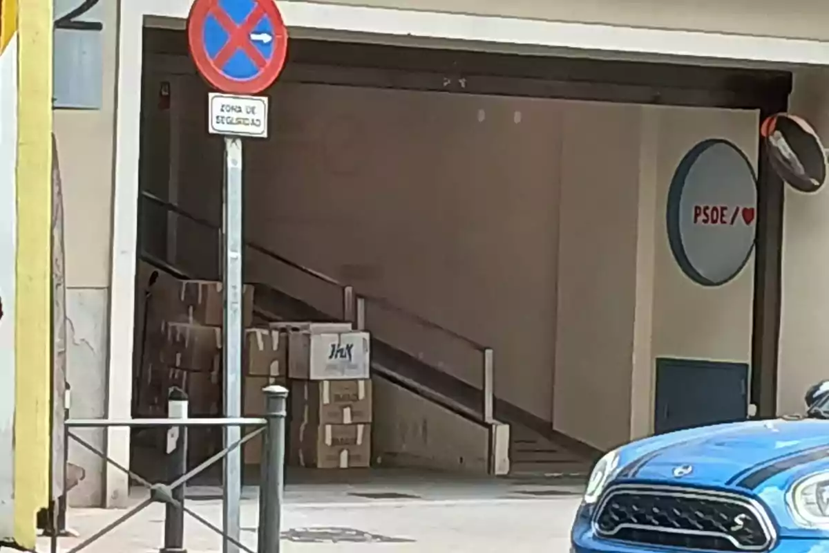 Un coche azul estacionado frente a una rampa con cajas apiladas al fondo, un letrero de no estacionar y un cartel de PSOE en la pared