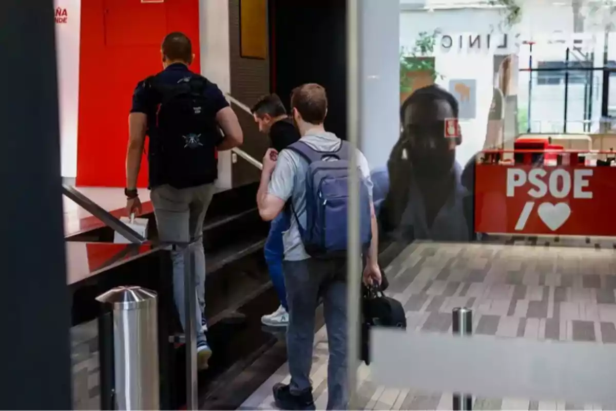Tres personas con mochilas suben unas escaleras dentro de un edificio moderno donde se observa un mostrador rojo con el logo de PSOE y un corazón Tres personas con mochilas suben unas escaleras dentro de un edificio moderno donde se observa un mostrador rojo con el logo de PSOE y un corazón