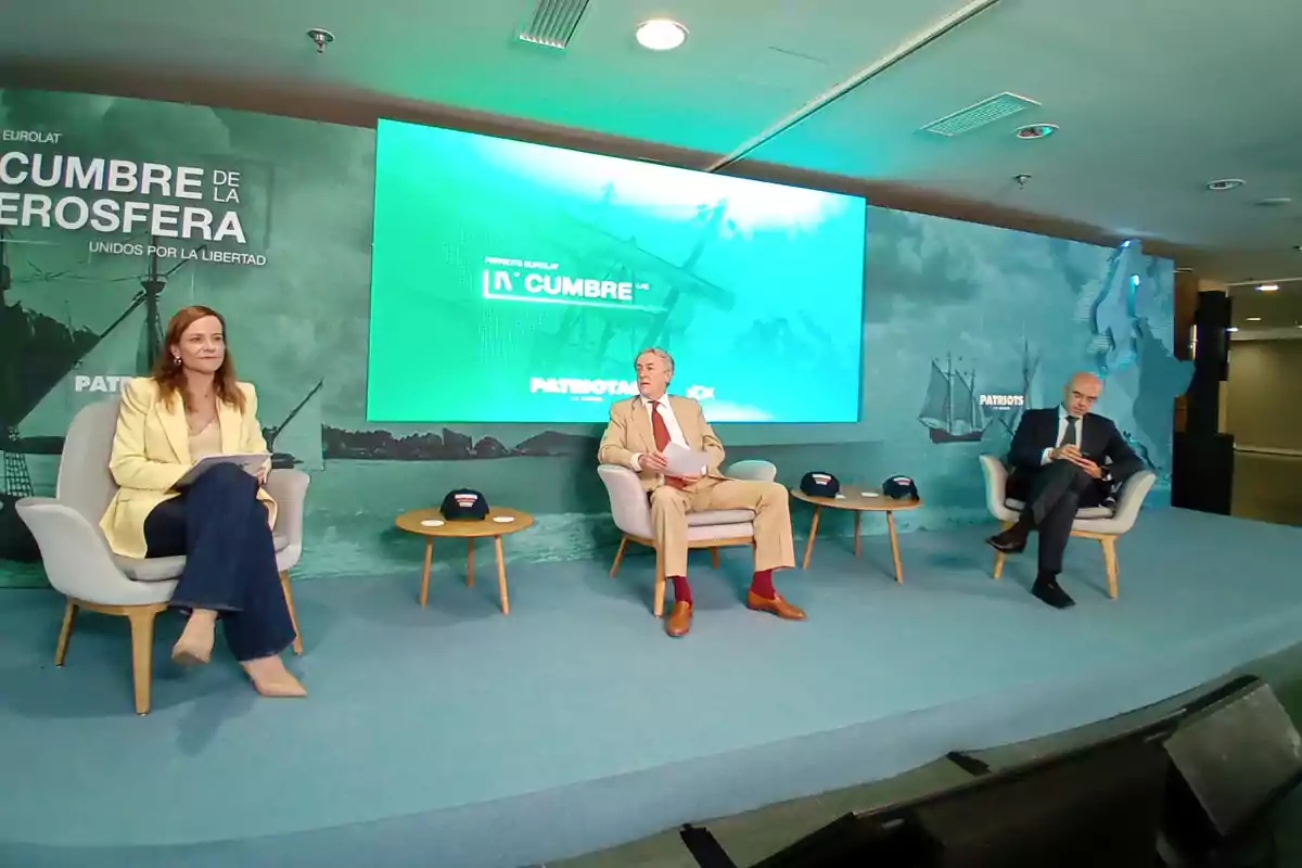 Tres personas sentadas en sillones sobre un escenario durante un evento con una pantalla grande de fondo que muestra el nombre de la cumbre y decoración relacionada con barcos antiguos