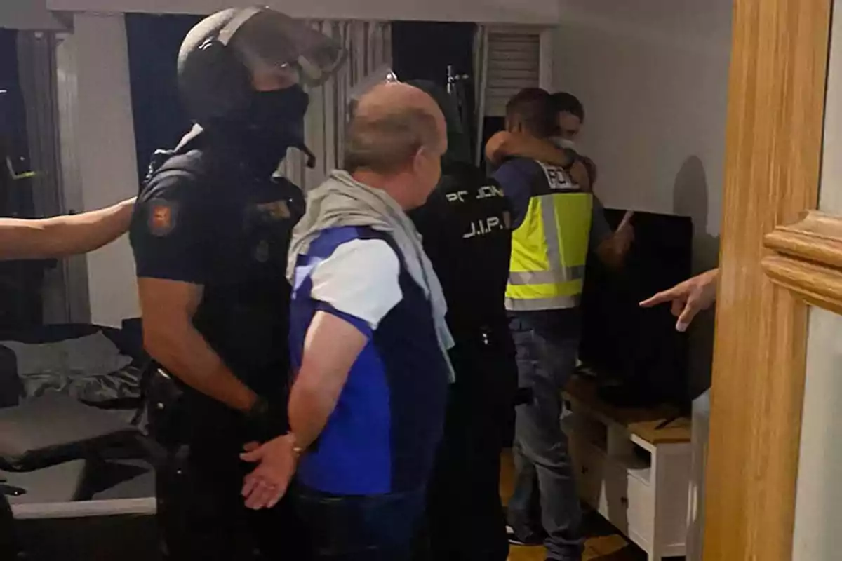 Varios policías realizan una detención dentro de una vivienda mientras una persona es esposada y otra es abrazada por un agente al fondo. Varios policías realizan una detención dentro de una vivienda mientras una persona es esposada y otra es abrazada por un agente al fondo.