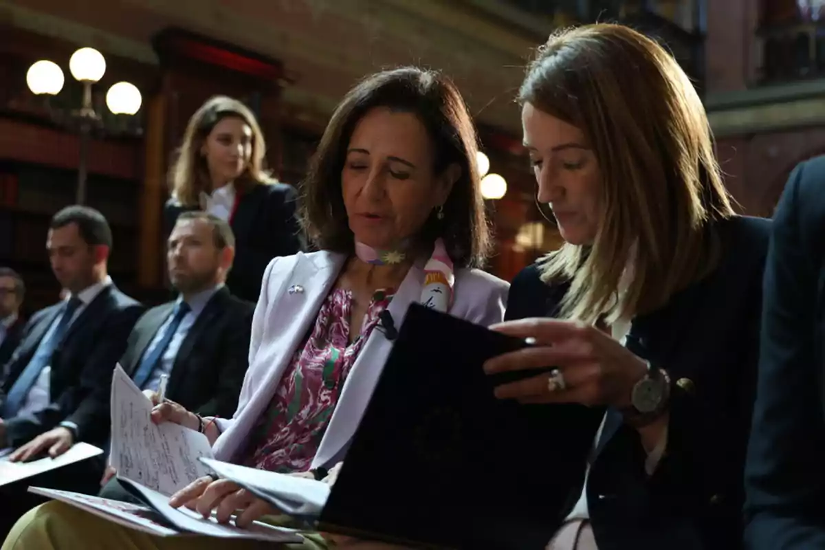 Dos mujeres revisan documentos sentadas en un evento formal rodeadas de otras personas en un ambiente elegante con estanterías y lámparas al fondo Dos mujeres revisan documentos sentadas en un evento formal rodeadas de otras personas en un ambiente elegante con estanterías y lámparas al fondo