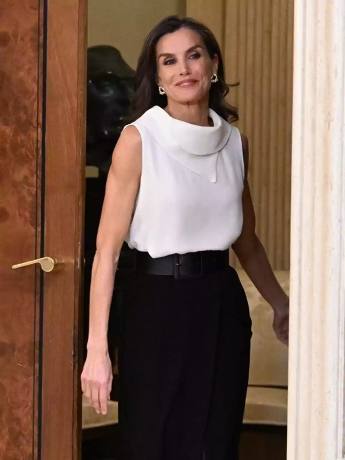 La reina Letizia de cabello oscuro y suelto entrando por una puerta, vestida con blusa blanca sin mangas y pantalón negro de talle alto con cinturón La reina Letizia de cabello oscuro y suelto entrando por una puerta, vestida con blusa blanca sin mangas y pantalón negro de talle alto con cinturón