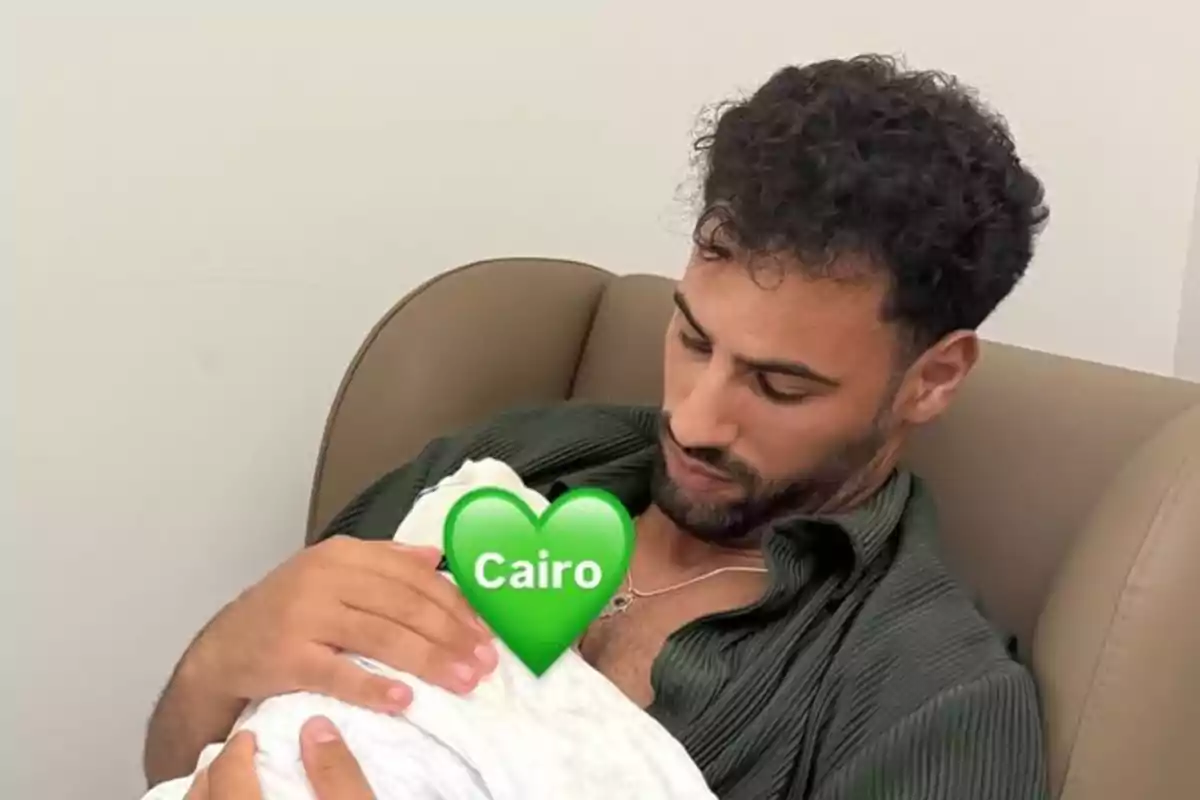 Asraf Beno sentado en un sillón sosteniendo a un bebé envuelto en una manta blanca con un emoji de corazón verde que dice Cairo cubriendo el rostro del bebé Asraf Beno sentado en un sillón sosteniendo a un bebé envuelto en una manta blanca con un emoji de corazón verde que dice Cairo cubriendo el rostro del bebé