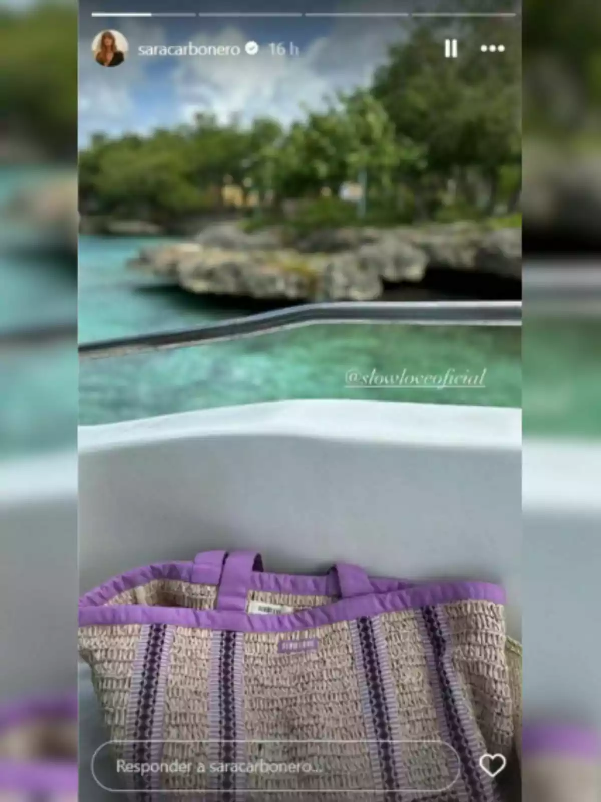 Bolso de rafia con detalles morados de Sara Carbonero apoyado en una barca blanca con agua turquesa y vegetación al fondo. Bolso de rafia con detalles morados de Sara Carbonero apoyado en una barca blanca con agua turquesa y vegetación al fondo.