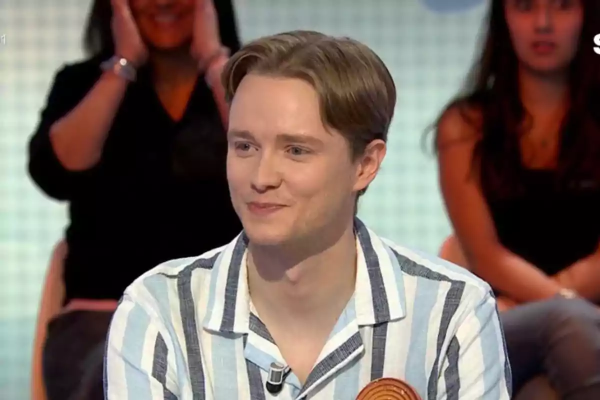 Manu de cabello rubio y camisa de rayas sonríe mientras participa en un programa de televisión con público al fondo Manu de cabello rubio y camisa de rayas sonríe mientras participa en un programa de televisión con público al fondo