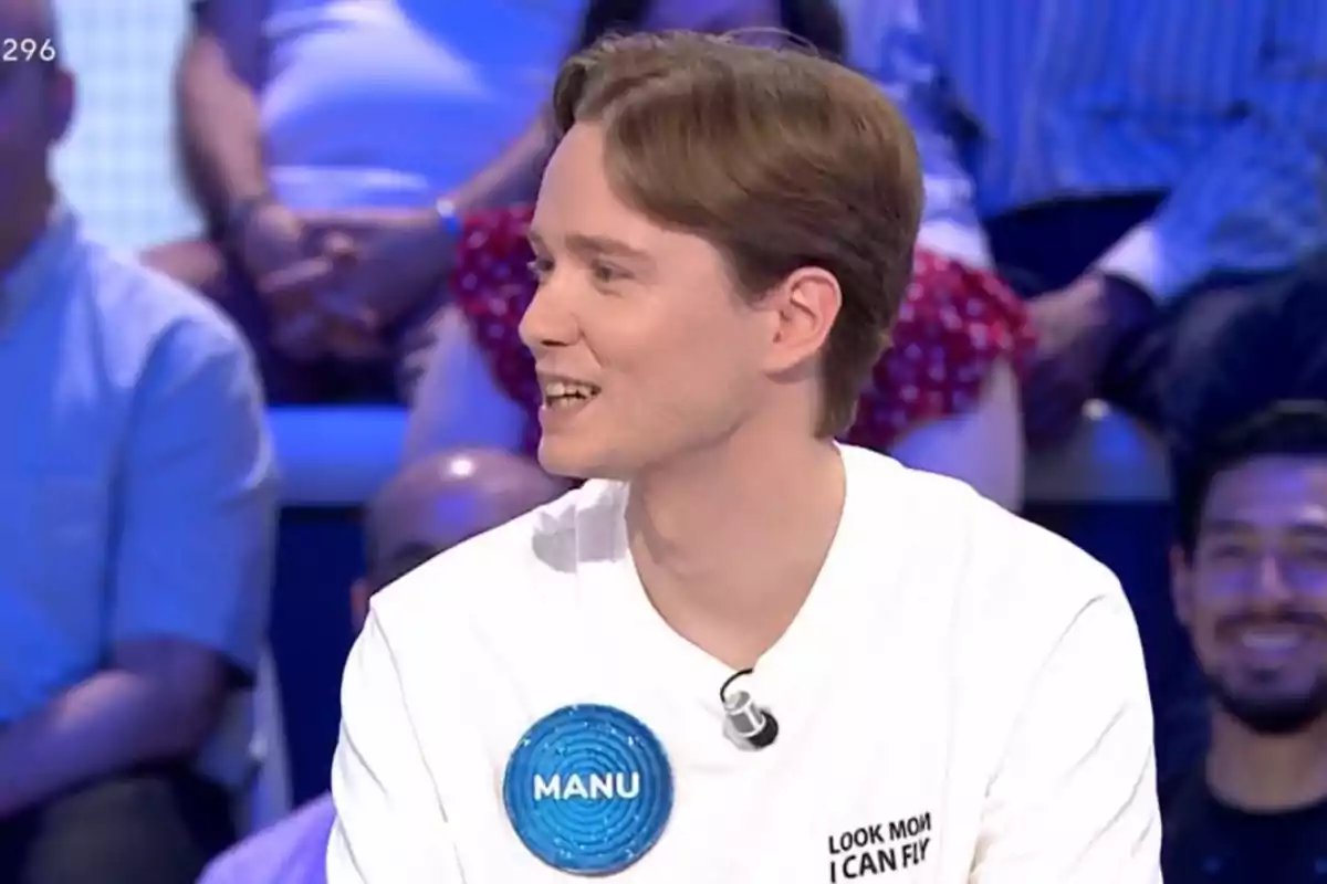 Manu con camiseta blanca y una placa azul que dice Manu participa en un programa de televisión con público al fondo Manu con camiseta blanca y una placa azul que dice Manu participa en un programa de televisión con público al fondo