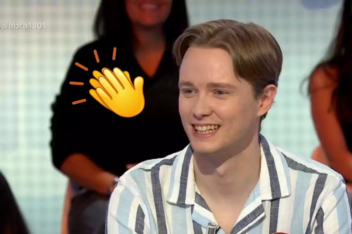 Manu sonriente con camisa de rayas azules y blancas en un programa de televisión con un emoji de aplausos al fondo