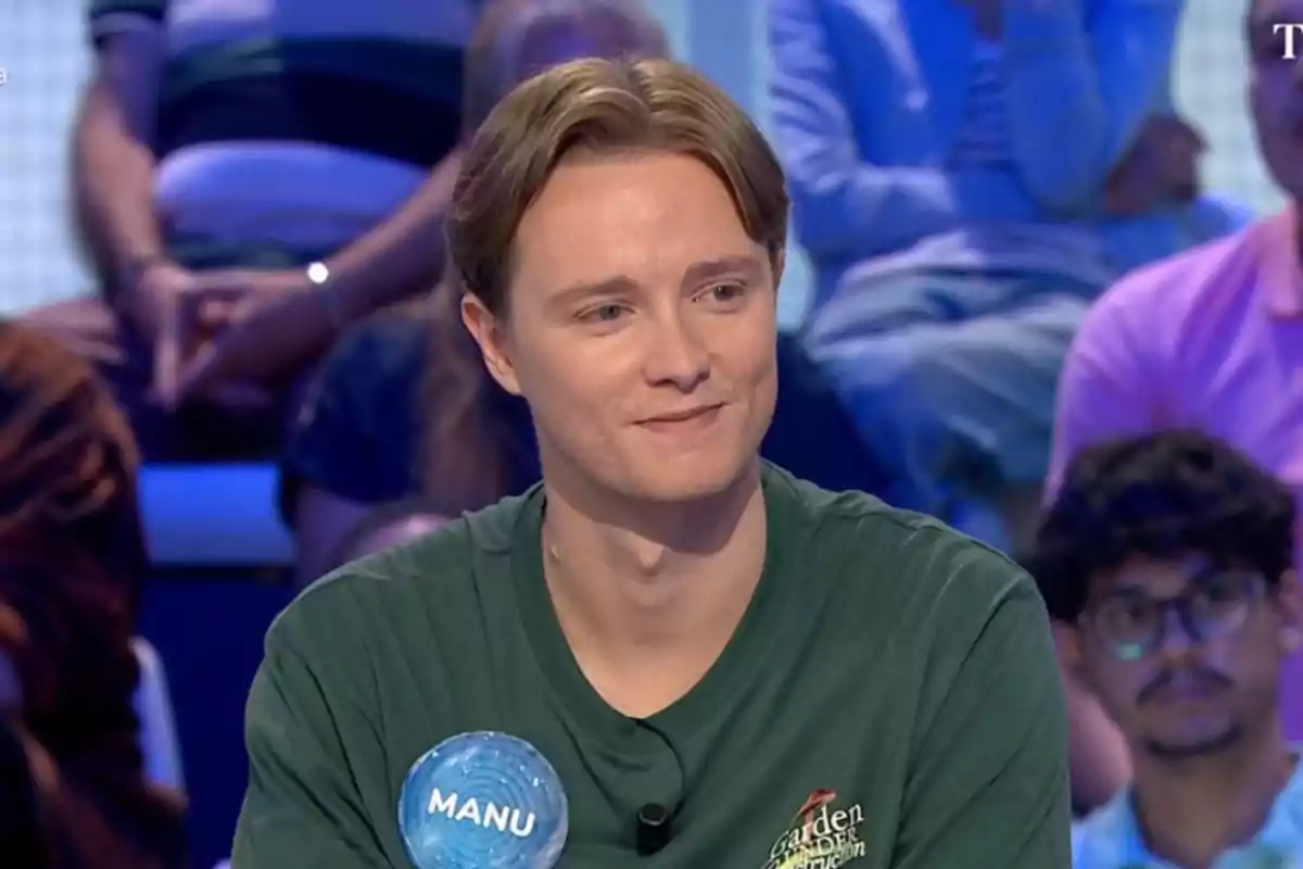 Manu con camiseta verde y una placa que dice Manu participa en un programa de televisión con público al fondo. Manu con camiseta verde y una placa que dice Manu participa en un programa de televisión con público al fondo.