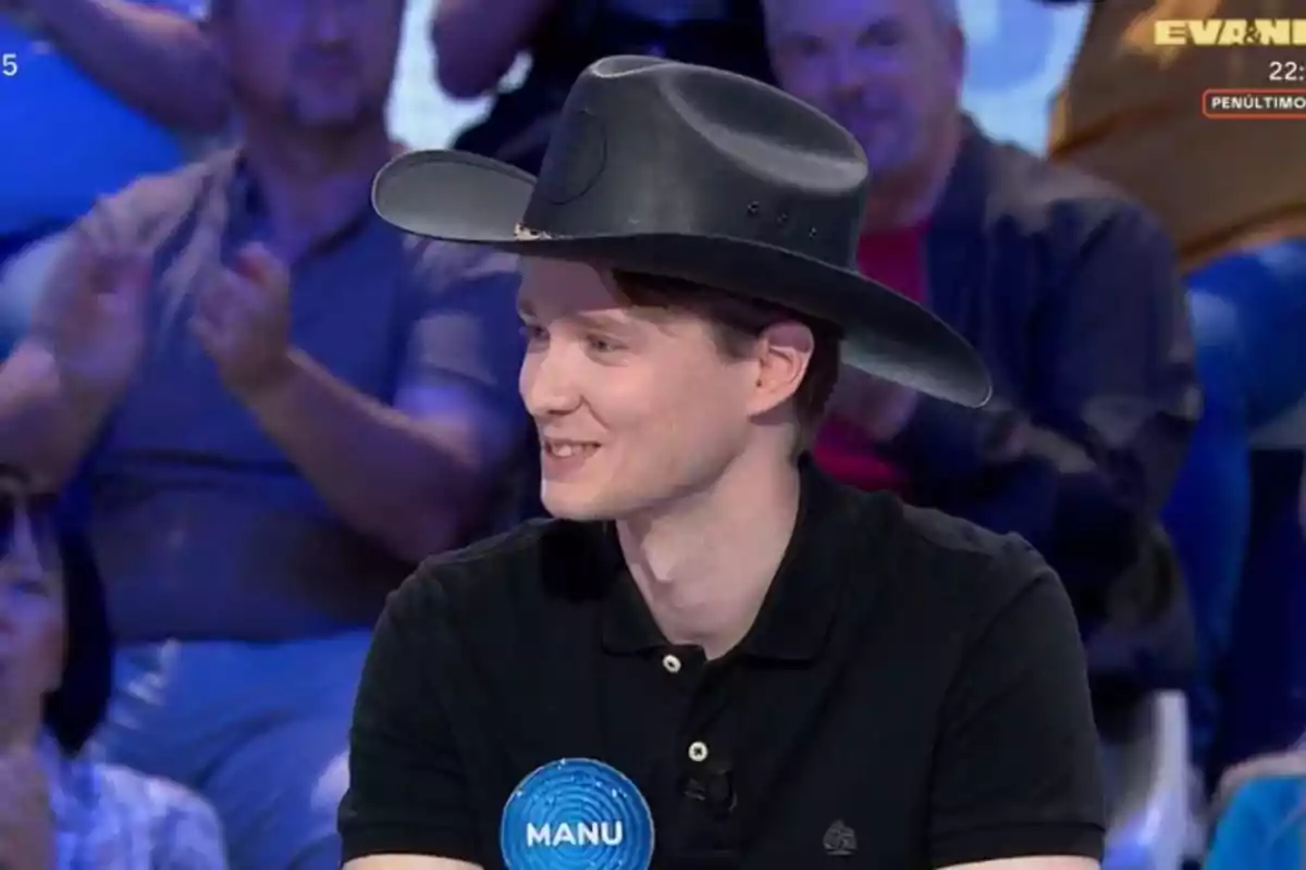 Manu con sombrero negro sonríe mientras participa en un programa de televisión, lleva una camiseta negra y una placa azul con el nombre Manu, el público aplaude en el fondo. Manu con sombrero negro sonríe mientras participa en un programa de televisión, lleva una camiseta negra y una placa azul con el nombre Manu, el público aplaude en el fondo.