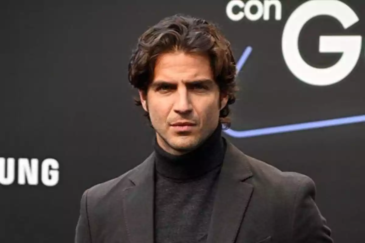 Maxi Iglesias de cabello ondulado y oscuro vistiendo un suéter de cuello alto negro y saco gris frente a un fondo oscuro con letras blancas Maxi Iglesias de cabello ondulado y oscuro vistiendo un suéter de cuello alto negro y saco gris frente a un fondo oscuro con letras blancas