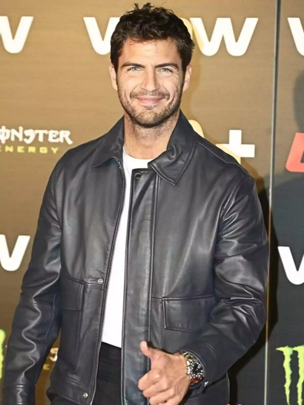 Maxi Iglesias de cabello corto y barba ligera vistiendo chaqueta de cuero negra y camiseta blanca posando sonriente en un evento frente a un fondo con logotipos. Maxi Iglesias de cabello corto y barba ligera vistiendo chaqueta de cuero negra y camiseta blanca posando sonriente en un evento frente a un fondo con logotipos.