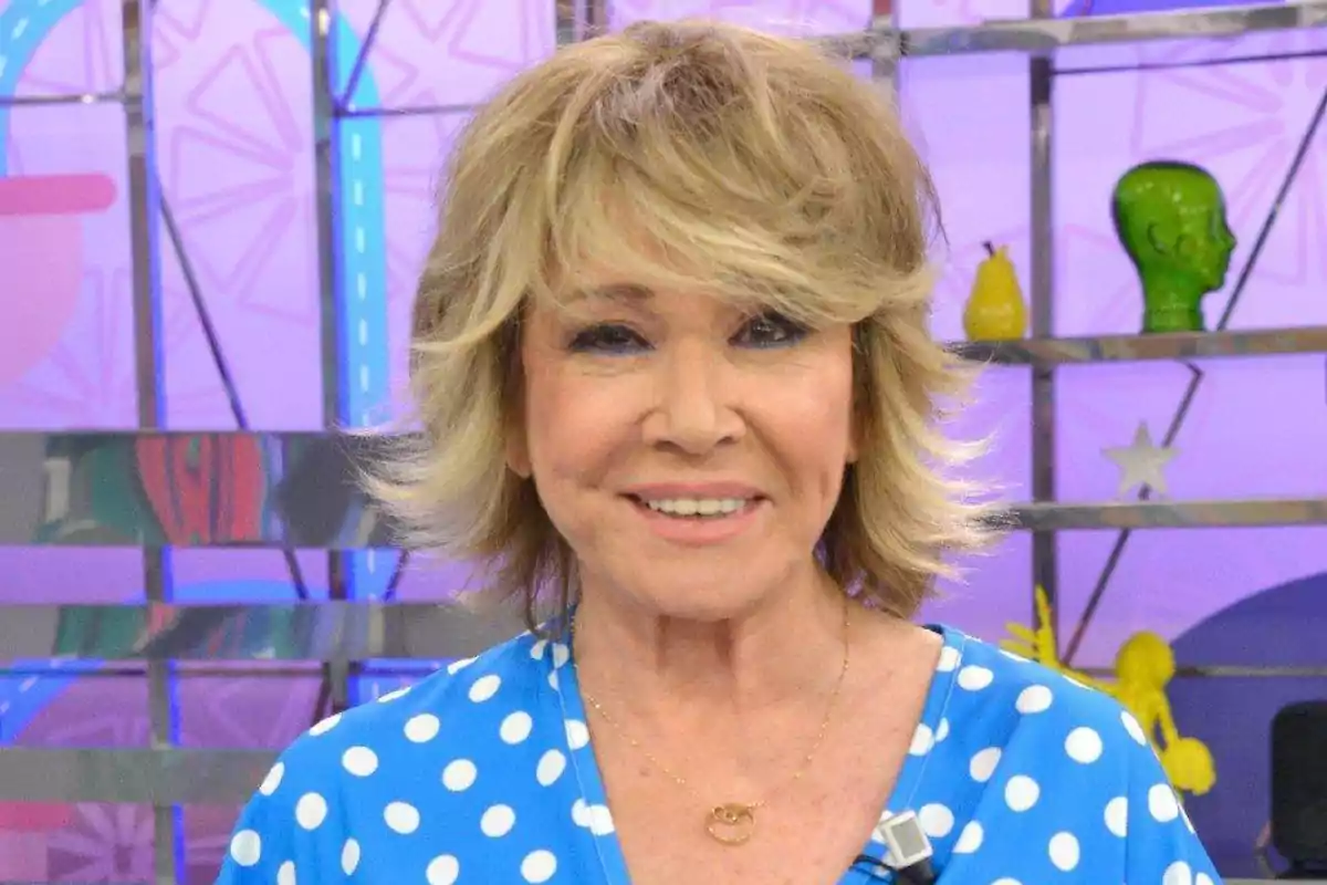 Mila Ximénez de cabello rubio corto con blusa azul de lunares blancos sonriendo en un set de televisión colorido Mila Ximénez de cabello rubio corto con blusa azul de lunares blancos sonriendo en un set de televisión colorido