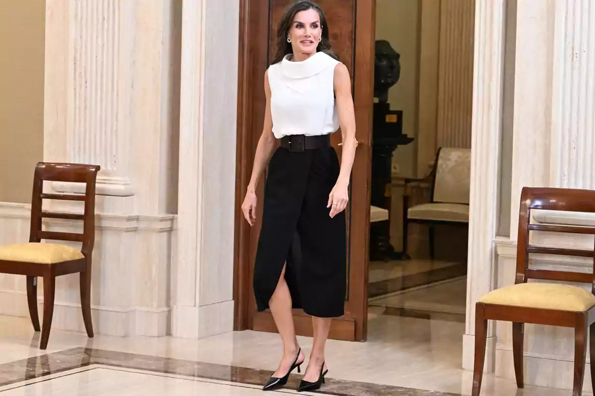 La reina Letizia de cabello largo y suelto vestida con blusa blanca sin mangas y falda negra midi con cinturón ancho, caminando en un elegante interior con sillas de madera y paredes de mármol. La reina Letizia de cabello largo y suelto vestida con blusa blanca sin mangas y falda negra midi con cinturón ancho, caminando en un elegante interior con sillas de madera y paredes de mármol.