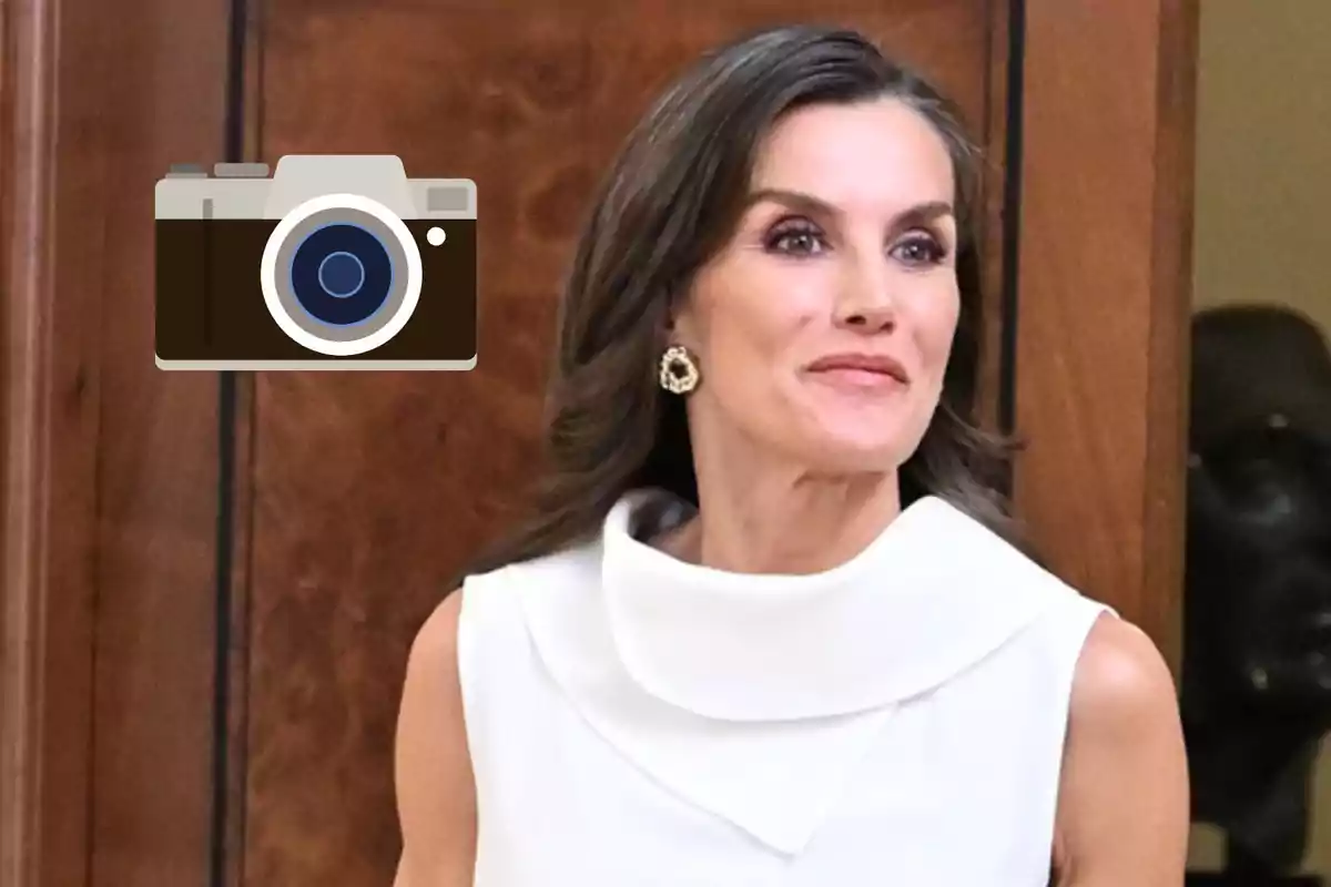 La reina Letizia de cabello castaño y vestido blanco posando frente a una pared de madera con un ícono de cámara superpuesto en la imagen