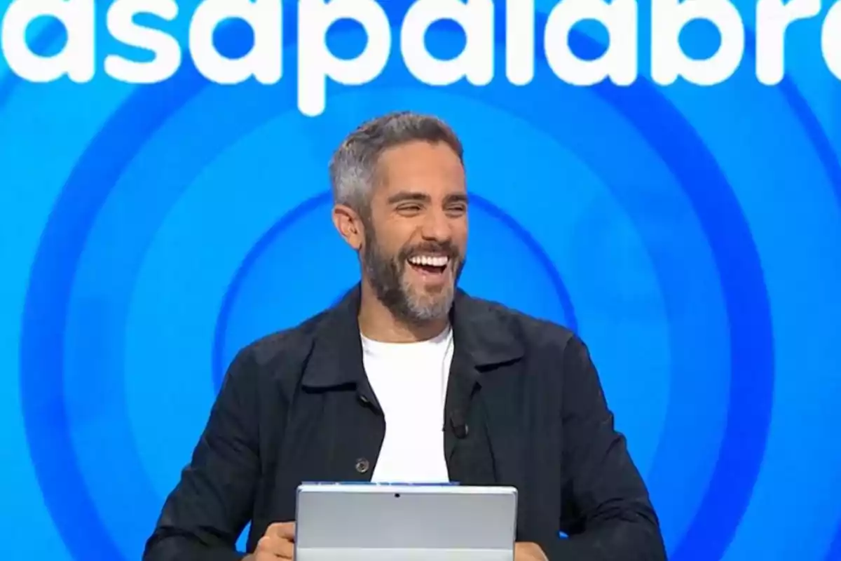 Roberto Leal de cabello corto y barba sonriendo mientras sostiene una tablet frente a un fondo azul con letras blancas Roberto Leal de cabello corto y barba sonriendo mientras sostiene una tablet frente a un fondo azul con letras blancas