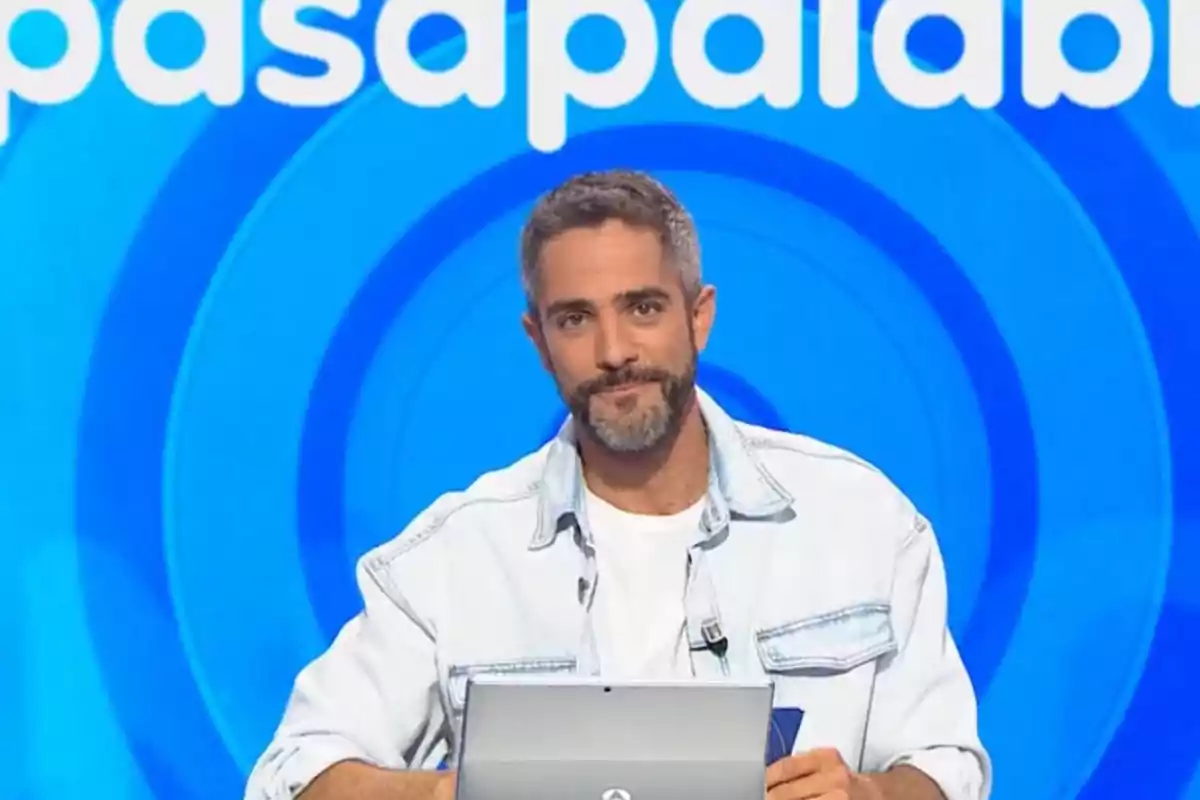 Roberto Leal de cabello corto y barba sentado frente a una computadora portátil con fondo azul y letras blancas de un programa de televisión Roberto Leal de cabello corto y barba sentado frente a una computadora portátil con fondo azul y letras blancas de un programa de televisión