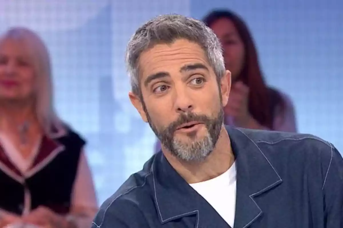 Roberto Leal de cabello y barba canosos con expresión de sorpresa en un set de televisión con personas desenfocadas al fondo Roberto Leal de cabello y barba canosos con expresión de sorpresa en un set de televisión con personas desenfocadas al fondo