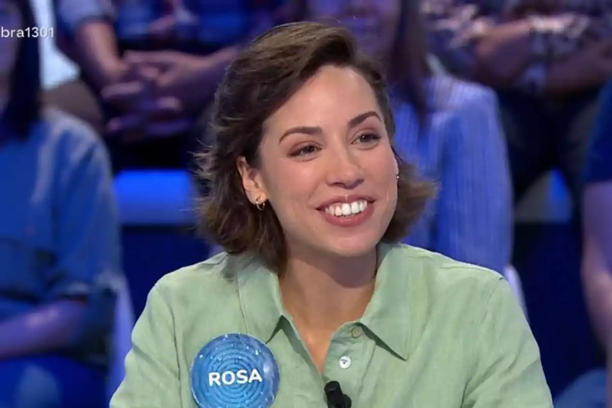 Rosa sonriente con cabello corto y camisa verde claro participando en un programa de televisión con una placa que dice Rosa Rosa sonriente con cabello corto y camisa verde claro participando en un programa de televisión con una placa que dice Rosa