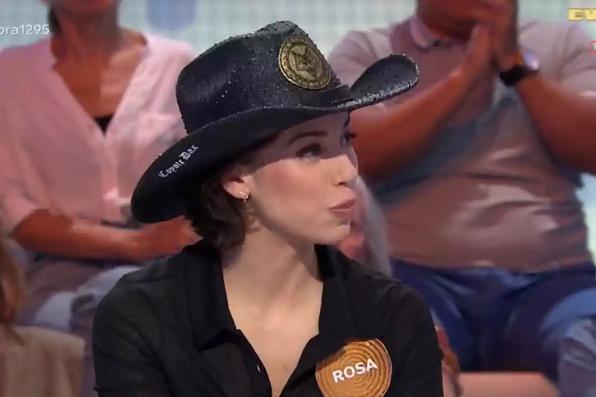 Rosa con sombrero negro y camisa oscura con una placa que dice Rosa participando en un programa de televisión con público de fondo Rosa con sombrero negro y camisa oscura con una placa que dice Rosa participando en un programa de televisión con público de fondo