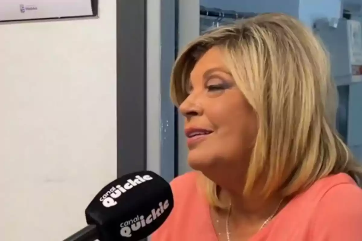 Terelu Campos rubia con blusa rosa hablando frente a un micrófono negro que tiene el logo de canal Quickie Terelu Campos rubia con blusa rosa hablando frente a un micrófono negro que tiene el logo de canal Quickie