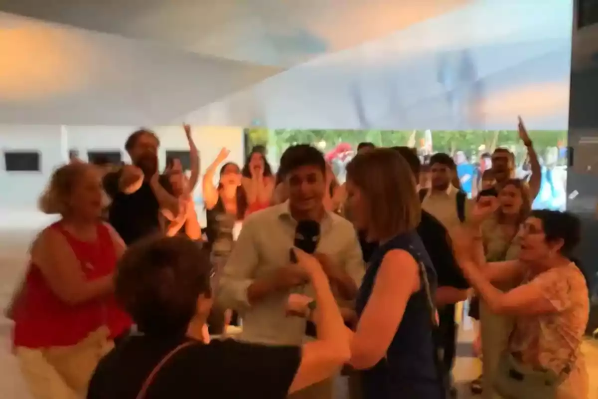 Un grupo de personas celebra animadamente en un espacio interior mientras una reportera entrevista a un hombre en el centro de la imagen