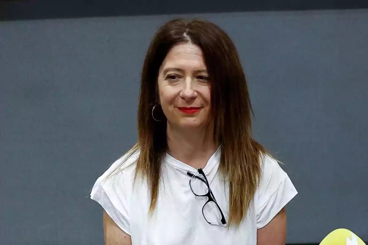 Mujer de cabello castaño lacio y largo con camiseta blanca y gafas colgadas en el cuello frente a un fondo gris Mujer de cabello castaño lacio y largo con camiseta blanca y gafas colgadas en el cuello frente a un fondo gris