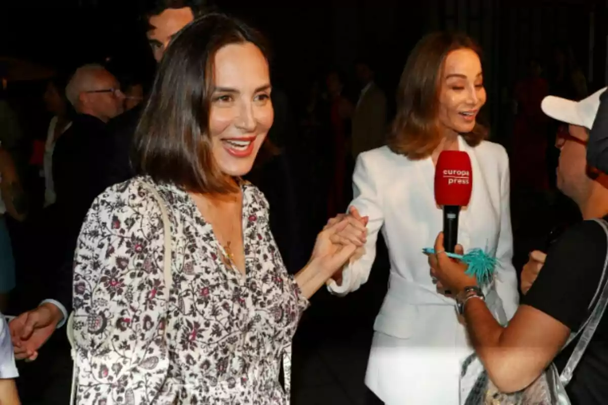 Isabel Preysler y Tamara Falcó sonrientes se saludan y se toman de la mano en un evento mientras una de ellas es entrevistada por un reportero que sostiene un micrófono rojo de Europa Press, rodeados de más personas en un ambiente nocturno. Isabel Preysler y Tamara Falcó sonrientes se saludan y se toman de la mano en un evento mientras una de ellas es entrevistada por un reportero que sostiene un micrófono rojo de Europa Press, rodeados de más personas en un ambiente nocturno.