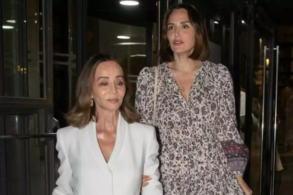Isabel Preysler y Tamara Falcó elegantemente vestidas salen de un edificio por una puerta de cristal, una lleva un conjunto blanco y la otra un vestido estampado. Isabel Preysler y Tamara Falcó elegantemente vestidas salen de un edificio por una puerta de cristal, una lleva un conjunto blanco y la otra un vestido estampado.