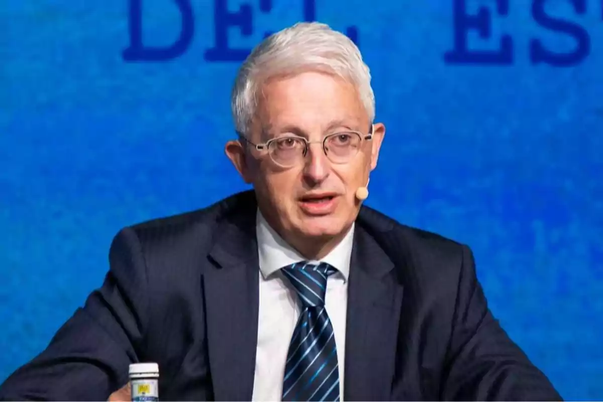 Hombre de cabello canoso y gafas hablando en un evento con micrófono de diadema y fondo azul Hombre de cabello canoso y gafas hablando en un evento con micrófono de diadema y fondo azul