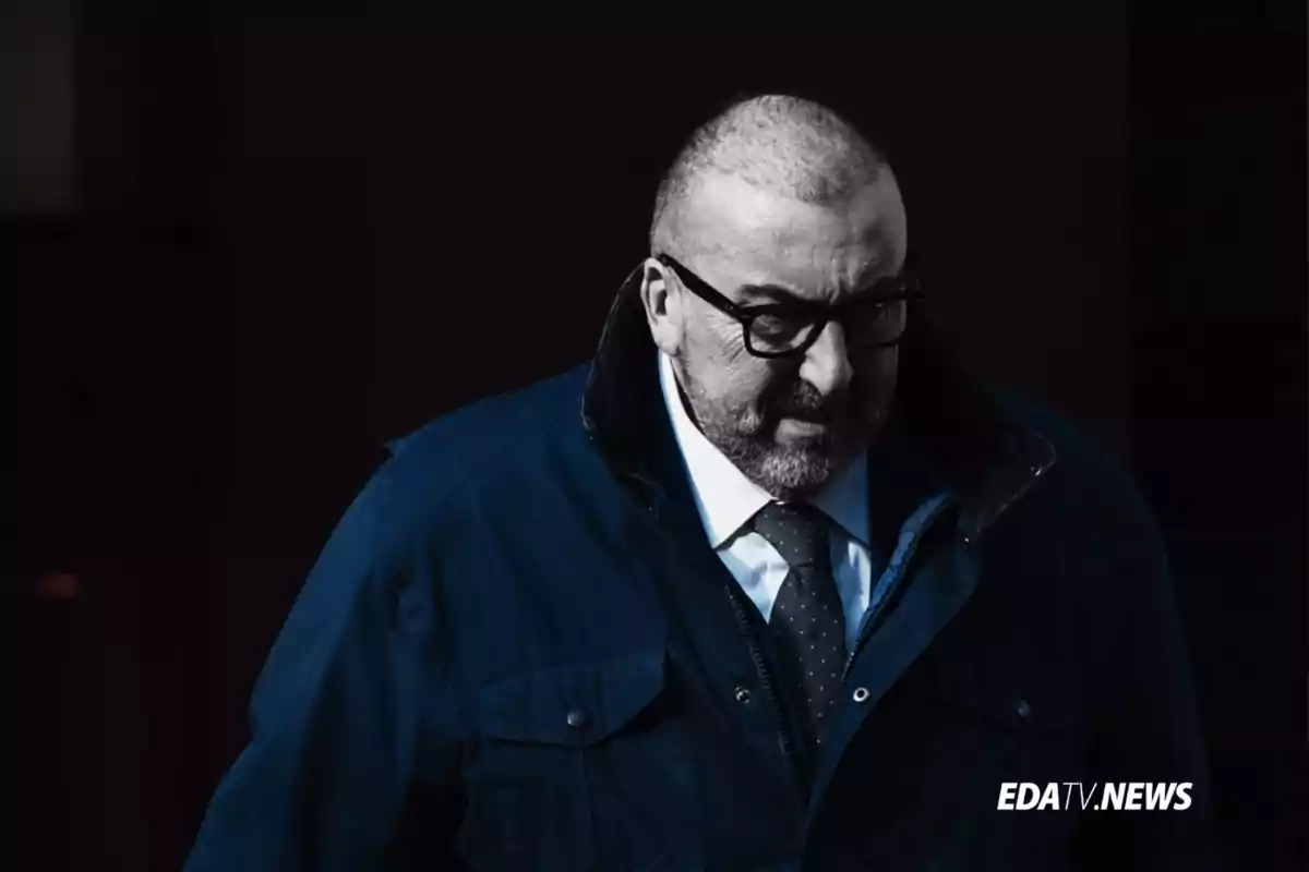 Hombre con gafas y barba vestido con abrigo azul y corbata en un entorno oscuro con el logo de EDATV NEWS en la esquina inferior derecha