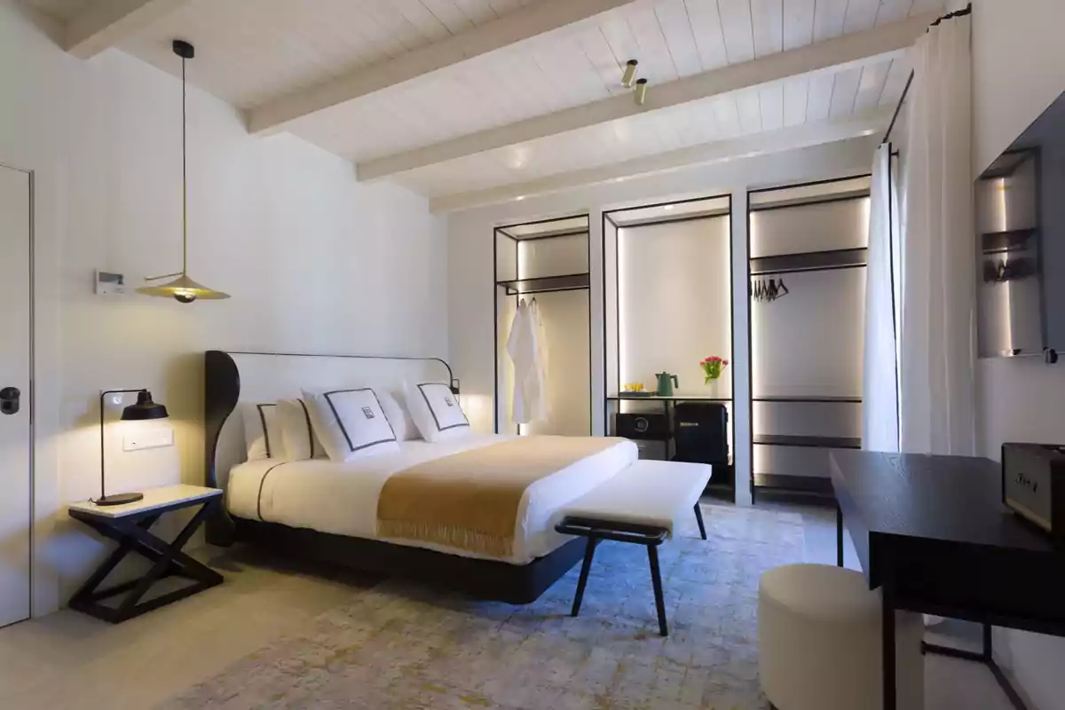 Habitación moderna de hotel con cama grande, ropa de cama blanca y beige, lámparas colgantes, armario abierto, escritorio negro y decoración minimalista