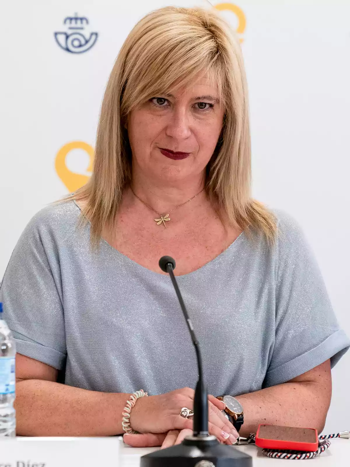 Mujer rubia sentada frente a un micrófono en una conferencia con fondo blanco y logotipos amarillos y azules Mujer rubia sentada frente a un micrófono en una conferencia con fondo blanco y logotipos amarillos y azules