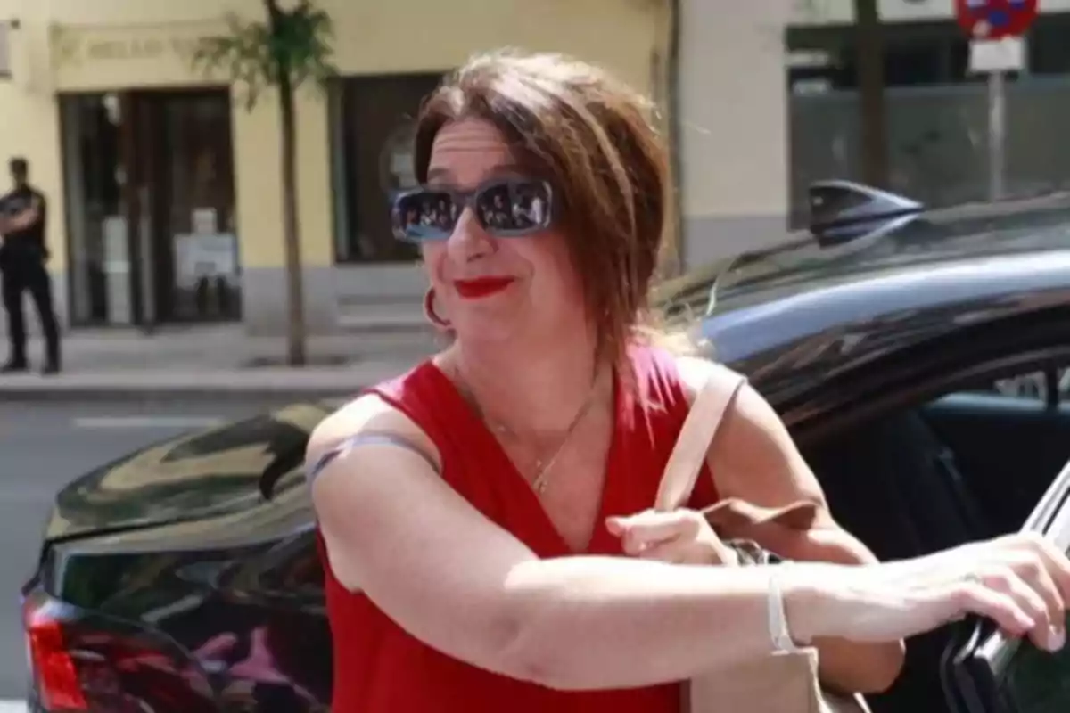 Mujer con gafas de sol y vestido rojo sonriendo mientras sale de un auto en una calle urbana Mujer con gafas de sol y vestido rojo sonriendo mientras sale de un auto en una calle urbana