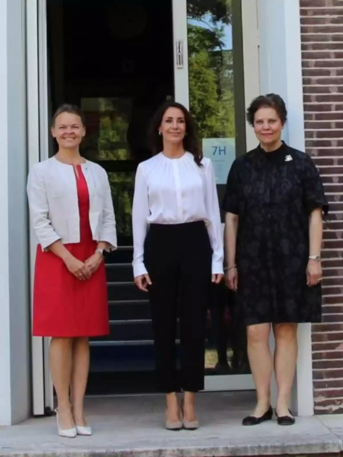 Three women stand posing in front of an entrance door, all dressed formally and smiling at the camera. Three women stand posing in front of an entrance door, all dressed formally and smiling at the camera.