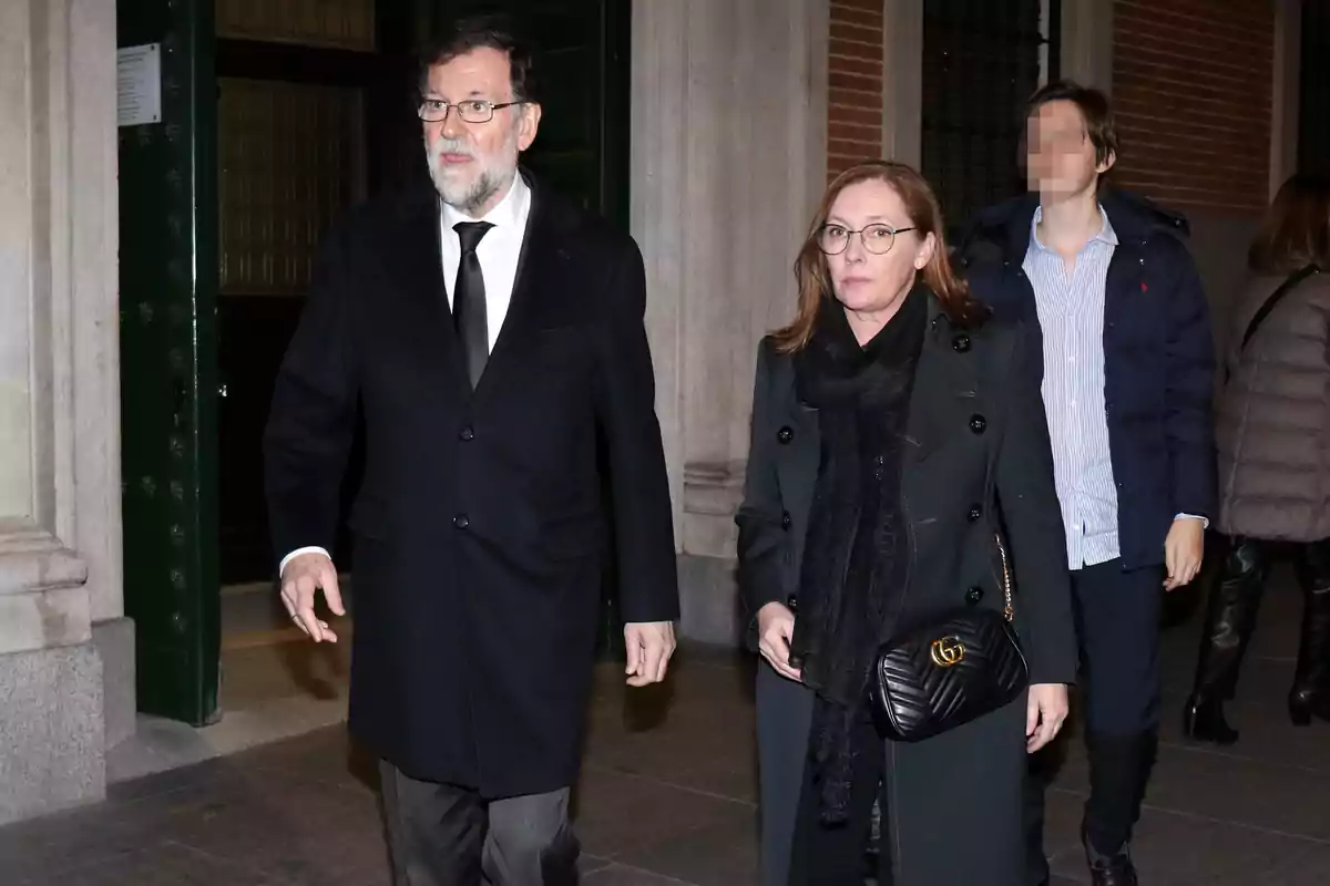 Mariano Rajoy y Elvira Fernández vestidos de negro caminan juntos por la calle, acompañados por una persona más al fondo. Mariano Rajoy y Elvira Fernández vestidos de negro caminan juntos por la calle, acompañados por una persona más al fondo.
